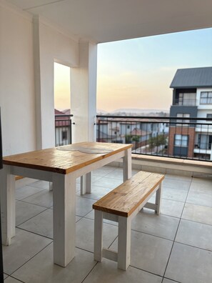 Ein Balkon mit Holztisch und Bank, weißen Wänden und Blick auf Gebäude.