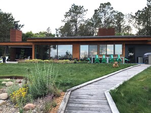 A modern house with a wooden deck, a garden with various plants, and a dog sitting on the grass. - Stunning 6-bedroom modern lake house perfect for families (Erhard)