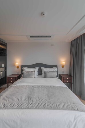A hotel room with a large bed, two bedside tables, a nightstand, a wall-mounted lamp, and a ceiling vent.