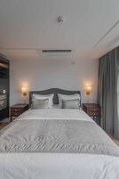A hotel room with a large bed, two bedside tables, a nightstand, a wall-mounted lamp, and a ceiling vent.