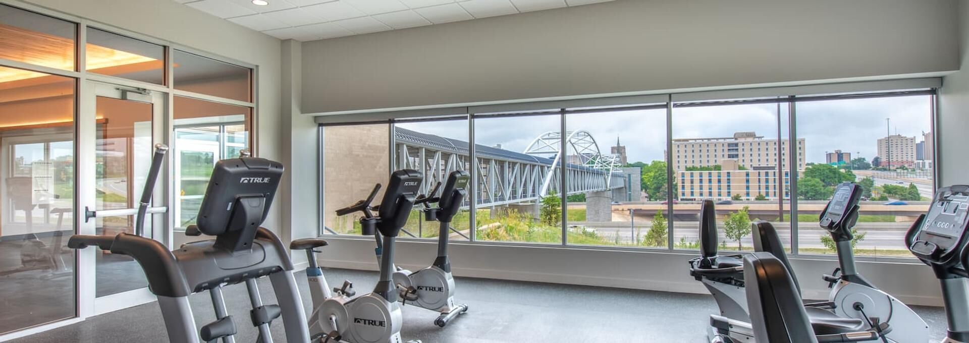 A gym with various exercise machines, large windows offering a view of a bridge and buildings.
