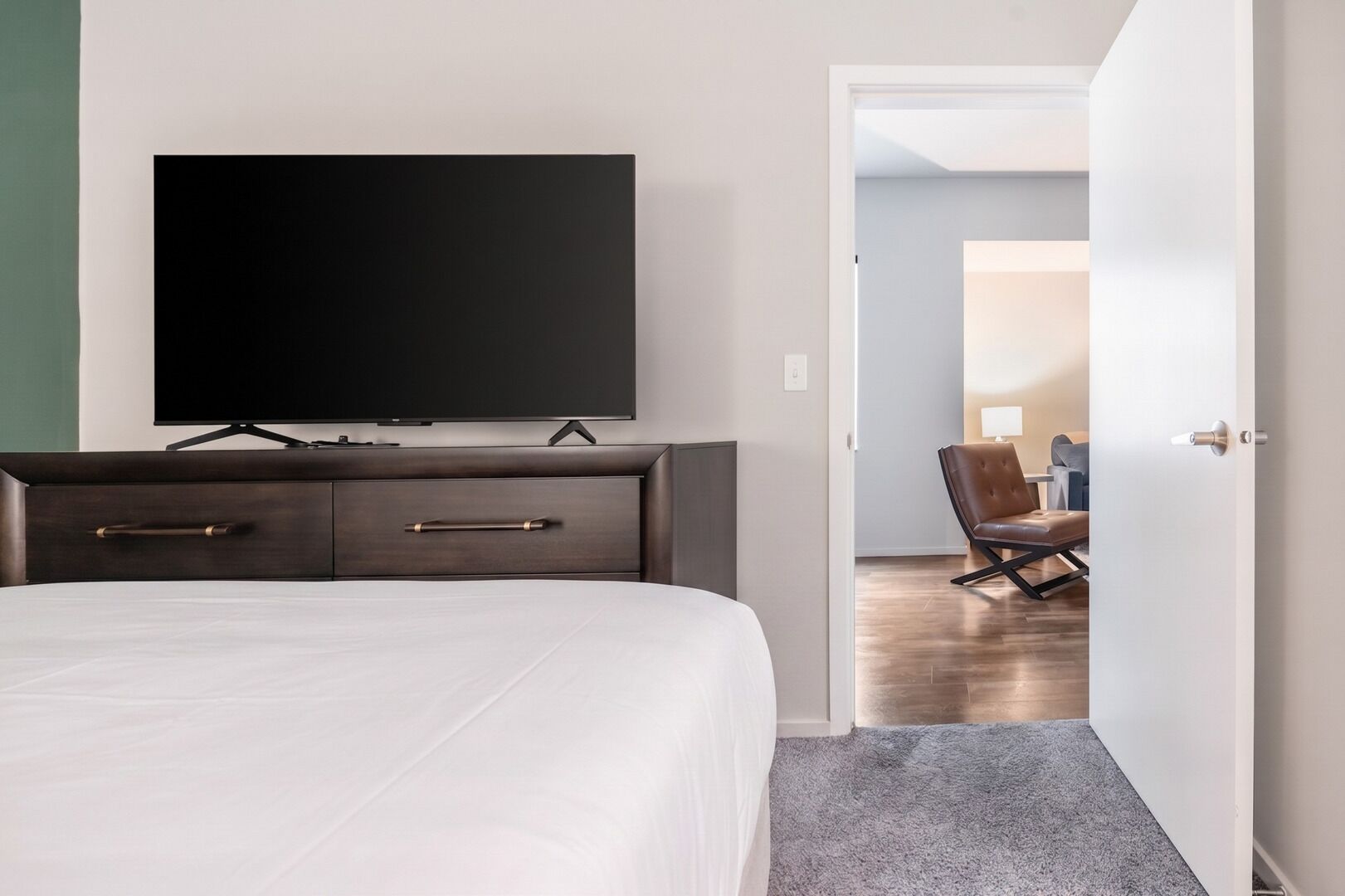A hotel room with a bed, a TV mounted on the wall, a desk with a chair, and a door leading to another room.