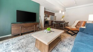 A modern living room with a flat-screen TV, wooden furniture, a coffee table, and a blue sofa. - Landing | Stunning 1BD, Gym, Yoga Studio (Omaha)