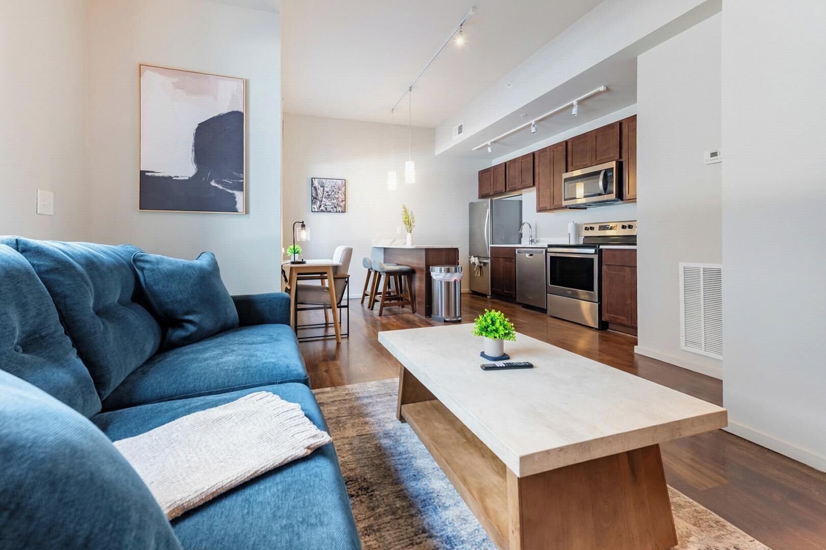 A modern living room with a blue sofa, a coffee table, and a kitchen area with wooden cabinets and stainless steel appliances.