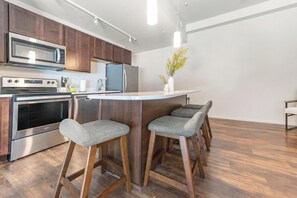 A modern kitchen with stainless steel appliances, wooden cabinets, and a bar stool seating area. - Landing | Modern 1BD, Yoga Studio, Gym (Omaha)