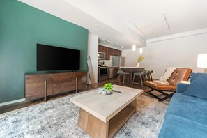 A modern living room with a flat-screen TV, wooden furniture, a dining area, and a kitchen. - Landing | Modern 1BD, Yoga Studio, Gym (Omaha)