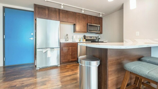 A modern kitchen with wooden cabinets, a stainless steel refrigerator, a microwave, and a bar counter with stools. - Landing | Stunning 1BD, Gym, Yoga Studio (Omaha)
