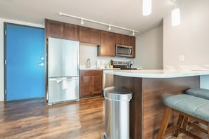 A modern kitchen with wooden cabinets, a stainless steel refrigerator, a microwave, and a bar counter with stools. - Landing | Modern 1BD, Yoga Studio, Gym (Omaha)