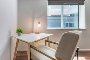 A room with a desk, chair, potted plant, and a window with a view of buildings. - Landing | Modern 1BD, Yoga Studio, Gym (Omaha)