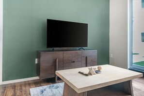 A modern living room with a flat-screen TV mounted on a dark wooden cabinet, a light-colored table with a remote control, and a blue patterned rug. - Landing | Incredible 1BD, Yoga Studio, Gym (Omaha)