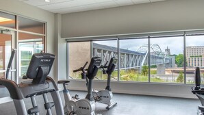 Een sportschool met diverse fitnessapparaten en grote ramen met uitzicht op een brug en gebouwen.