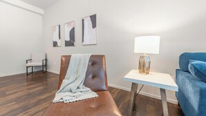 Een kamer met een bruine leren bank, een blauwe slaapbank, een klein wit tafeltje met een lamp en drie abstracte schilderijen aan de muur.