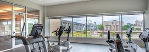 A gym with multiple exercise machines, large windows offering a view of a bridge and buildings. - Landing | Chic 2BD, Yoga Studio, Gym (Omaha)