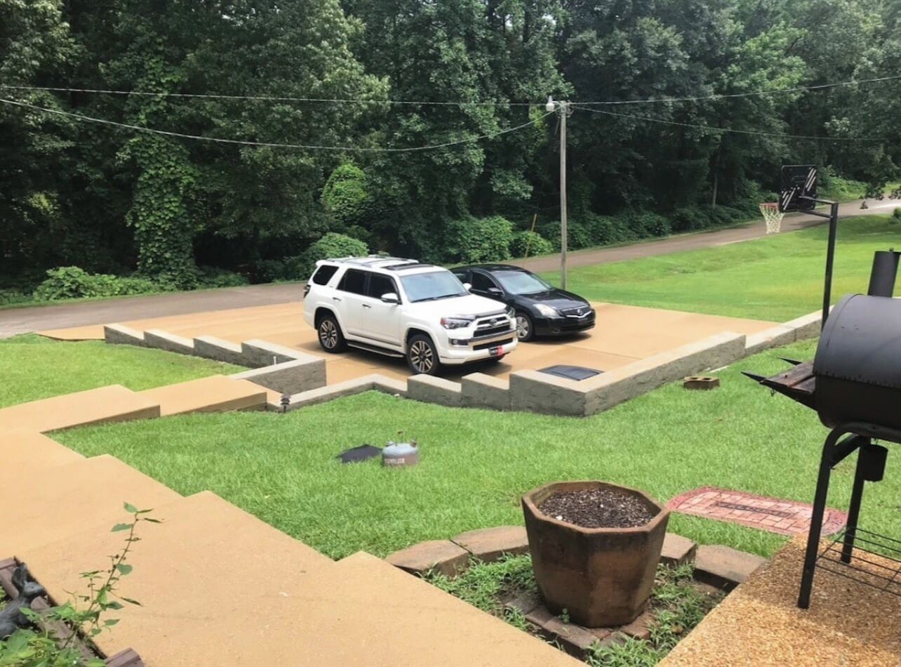 A backyard with a barbecue grill, a white SUV, and a black car parked on a paved area.