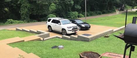 A backyard with a barbecue grill, a white SUV, and a black car parked on a paved area.