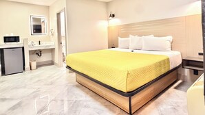 Habitación de hotel con cama, mesita de noche, escritorio con silla, espejo y refrigerador pequeño.