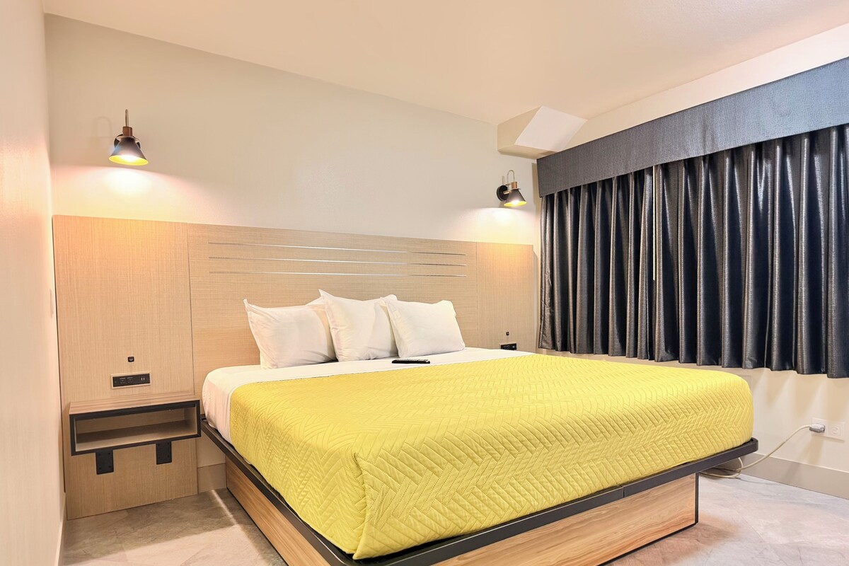 Quarto de hotel moderno com cabeceira de madeira, colcha de cama acolchoada amarela, duas luminárias de mesa e uma janela grande com cortinas escuras.