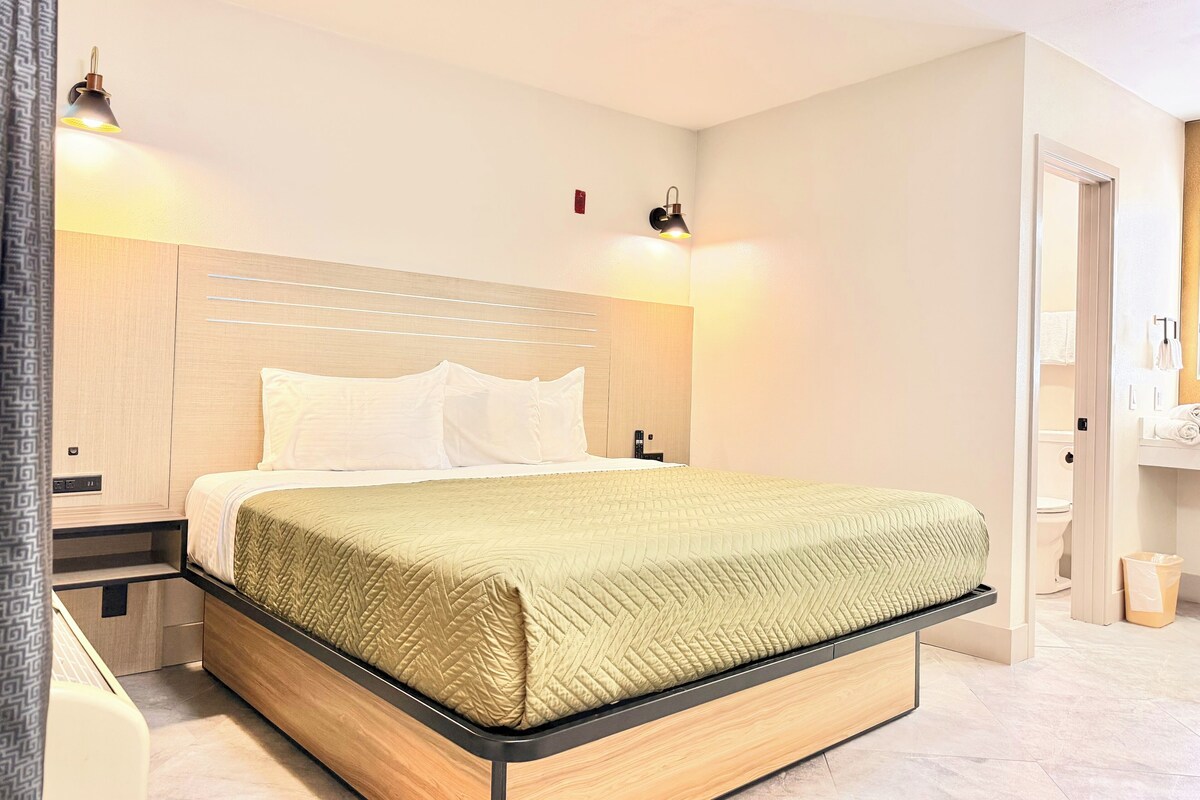 Una habitación de hotel moderna con cama, mesitas de noche y un baño visible a través de una puerta abierta.