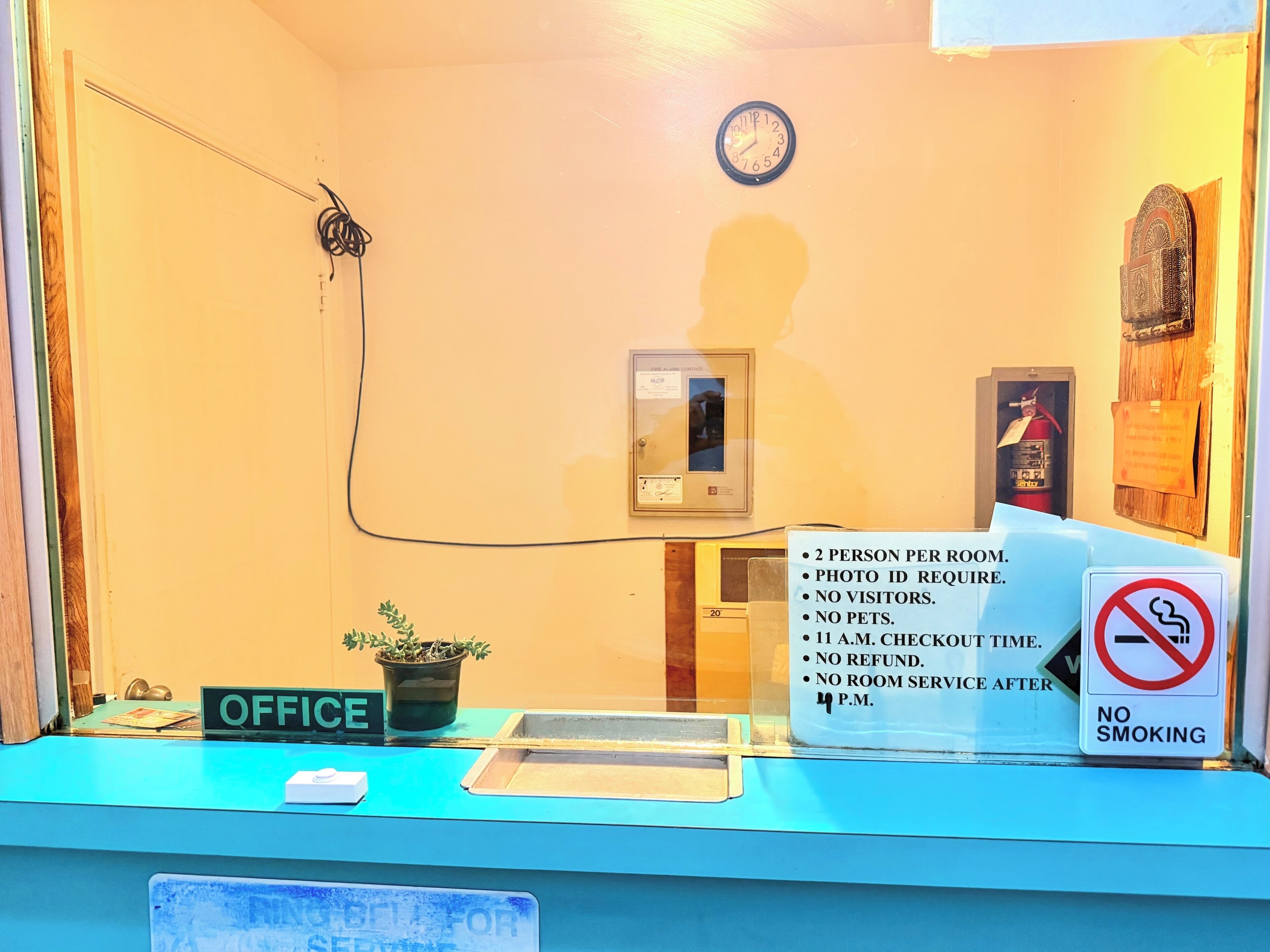 A reception desk with a sign indicating a 2 person per room policy, photo ID requirement, no visitors, no pets, 11 a.m. checkout time, no refund, and no room service after 4 p.m.