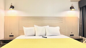 Habitación de hotel con cabecera de madera, dos mesitas de noche con lámparas, cama con ropa blanca y colcha amarilla.