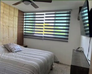A bedroom with a bed, a ceiling fan, a window with blinds, and a TV mounted on the wall. - 2-bedroom apartment in welcoming Mazatlán with AC for comfort (Mazatlán)