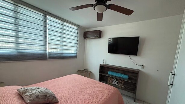 A bedroom with a bed, a ceiling fan, a TV mounted on the wall, and a small table. - 2-bedroom apartment in welcoming Mazatlán with AC for comfort (Mazatlán)