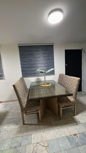 A dining area with a glass table and patterned chairs, a potted plant, and a window with blinds. - 2-bedroom apartment in welcoming Mazatlán with AC for comfort (Mazatlán)