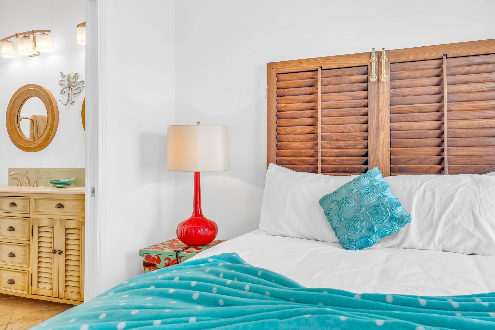 A bedroom with a wooden headboard, a red floor lamp, a bed with white and blue bedding, a blue polka dot blanket, and a small bedside table with a green patterned box.