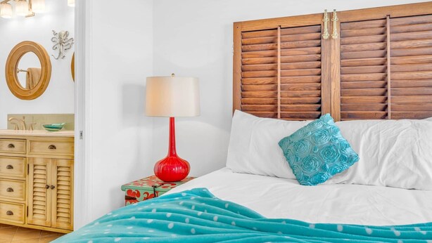 A bedroom with a wooden headboard, a red floor lamp, a bed with white and blue bedding, a blue polka dot blanket, and a small bedside table with a green patterned box. - Pet-Friendly Boater’s Retreat- Pool, Dock & Kayaks (Marathon)