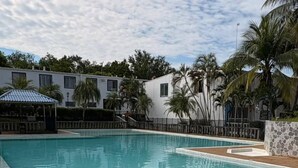 A swimming pool with a tiled edge, surrounded by palm trees and a building with multiple windows.