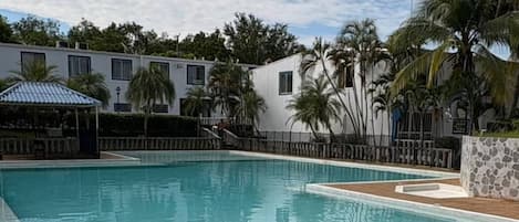 Una piscina con borde de azulejos, rodeada de palmeras y un edificio con varias ventanas.