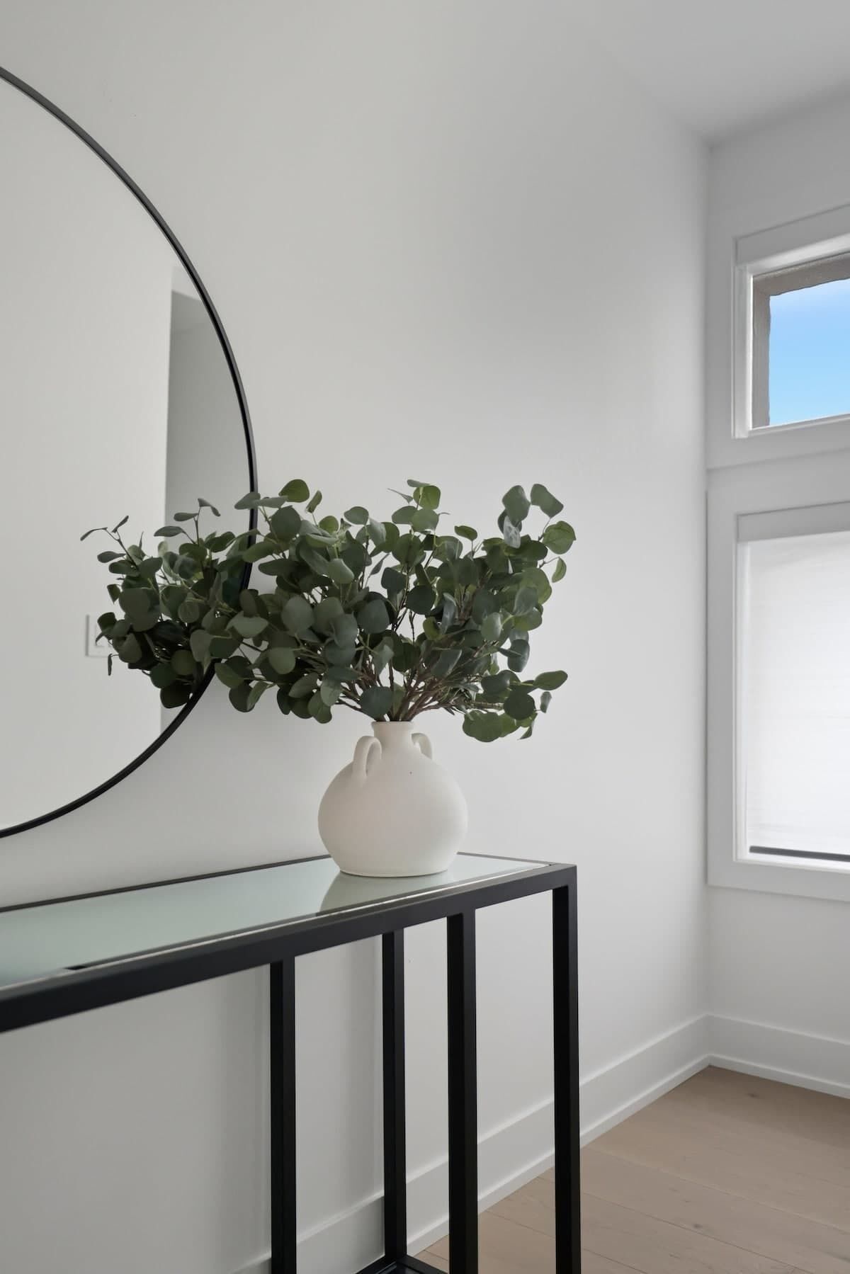 Un vase blanc orné de feuillage vert, posé sur une console noire, dans une pièce aux murs blancs et dotée d’un grand miroir.