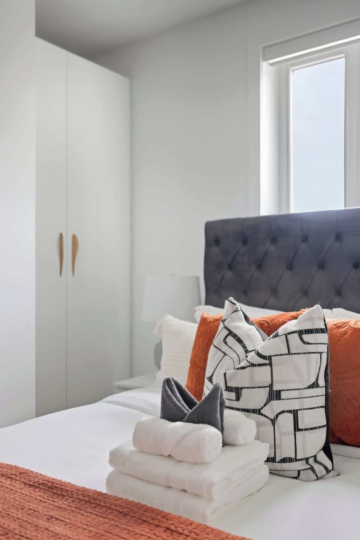 Una cama bien hecha con almohadas y una toalla doblada, un armario de dos puertas y una ventana con luz natural.