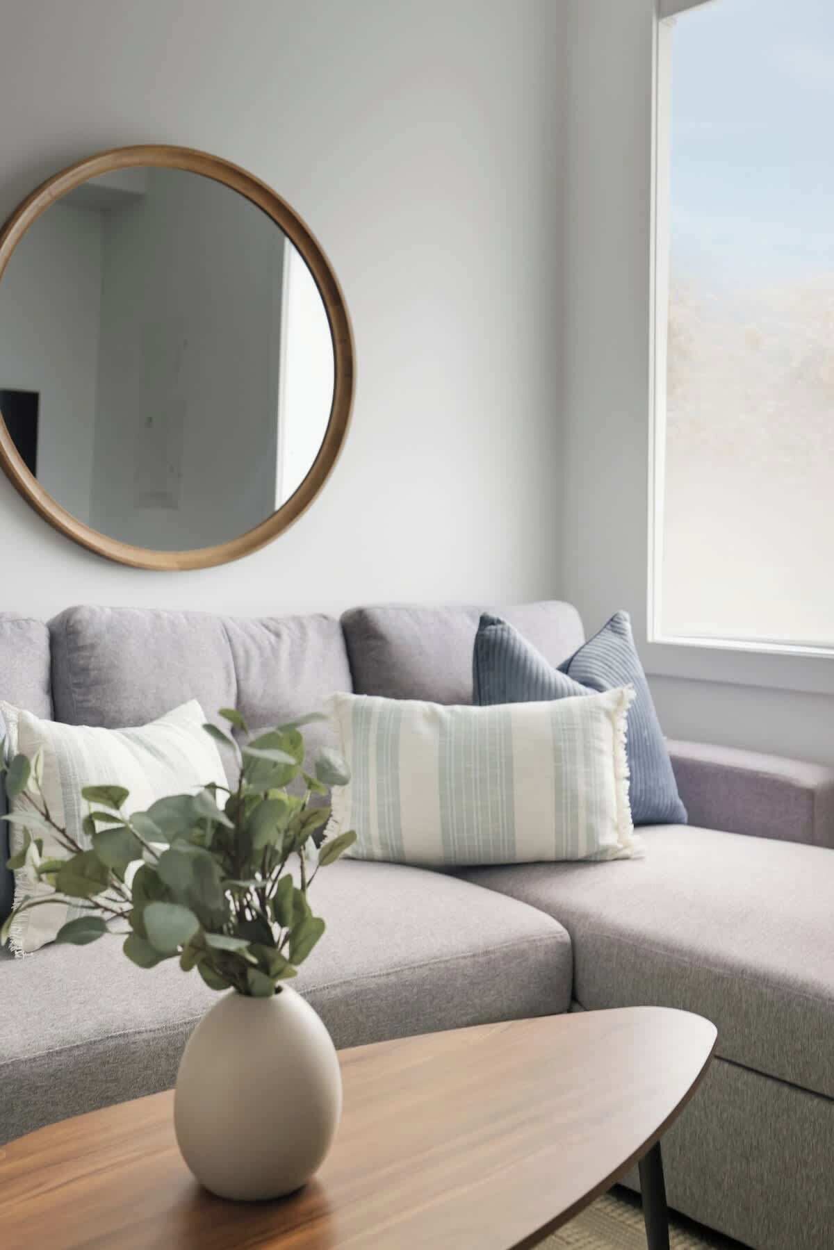 A modern living room with a grey sofa, a round mirror, and a wooden coffee table with a plant.