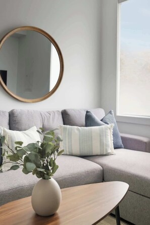 A modern living room with a grey sofa, a round mirror, and a wooden coffee table with a plant. - Roncesvalles Charm Stylish 1BR Near High Park! | Parking Available (Toronto)