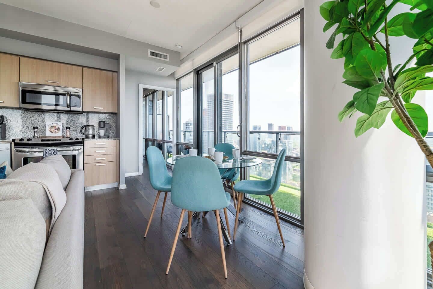 Une cuisine moderne dotée d’un four et d’un micro-ondes encastrés, un coin repas avec une table en verre et des chaises bleues, et de grandes fenêtres offrant une vue sur la ville.