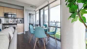 Une cuisine moderne dotée d’un four et d’un micro-ondes encastrés, un coin repas avec une table en verre et des chaises bleues, et de grandes fenêtres offrant une vue sur la ville.