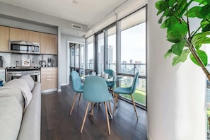 A modern kitchen with a built-in oven and microwave, a dining area with a glass table and blue chairs, and large windows offering a city view.