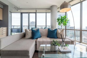 A modern living room with a sectional sofa, a glass coffee table, and a large window offering a city view. - Premier Sub-Penthouse w Panoramic Views N Parking! (Toronto)