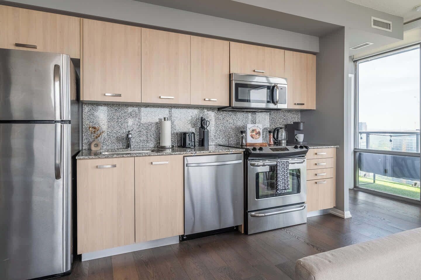 A modern kitchen with stainless steel appliances, wooden cabinets, and a large window offering a view.