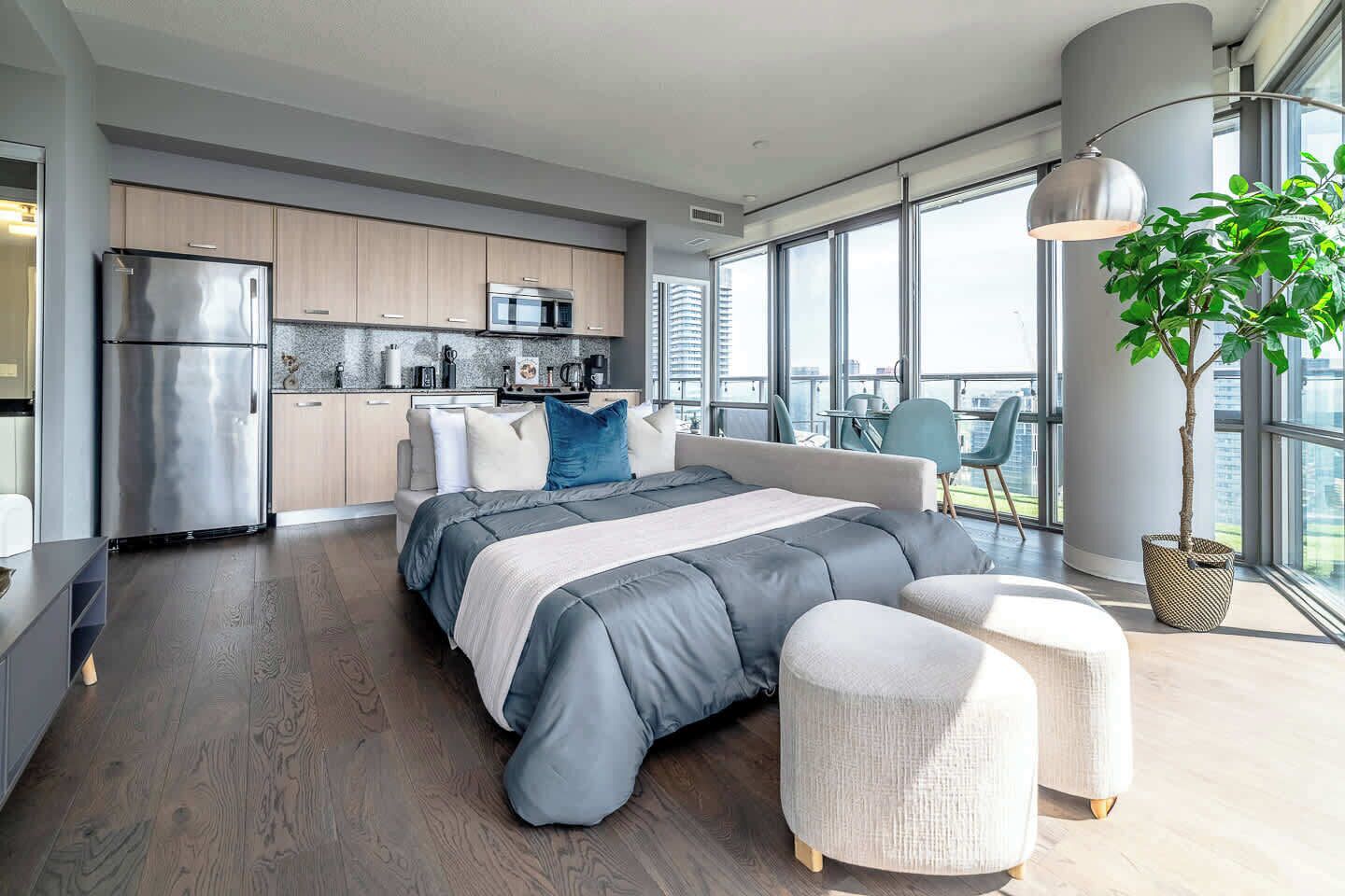 A modern bedroom with a large bed, a view of the city, and a kitchenette in the background.