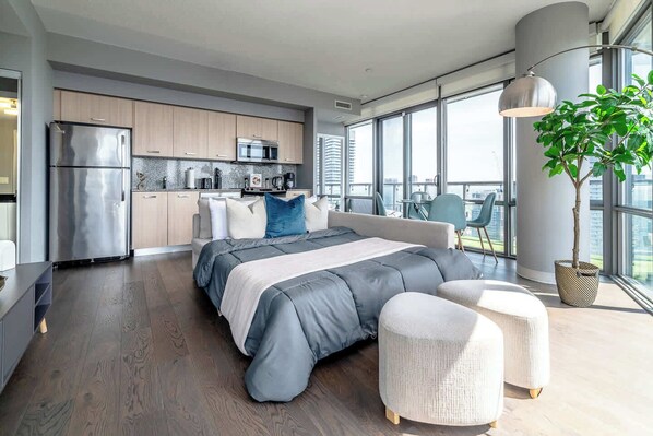 A modern bedroom with a large bed, a view of the city, and a kitchenette in the background.