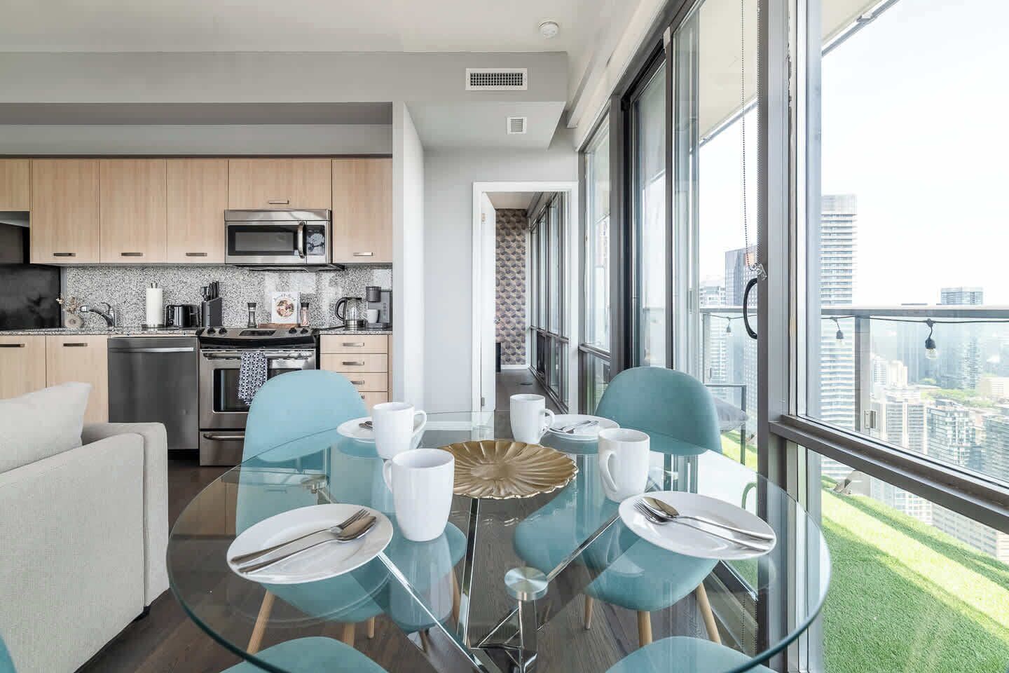 A modern kitchen with stainless steel appliances, a dining area with glass tables and chairs, and a view of a cityscape through large windows.