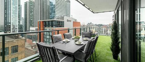 Un balcon avec une table et des chaises pour prendre les repas, une petite plante en pot et une vue sur un paysage urbain avec de hauts immeubles.