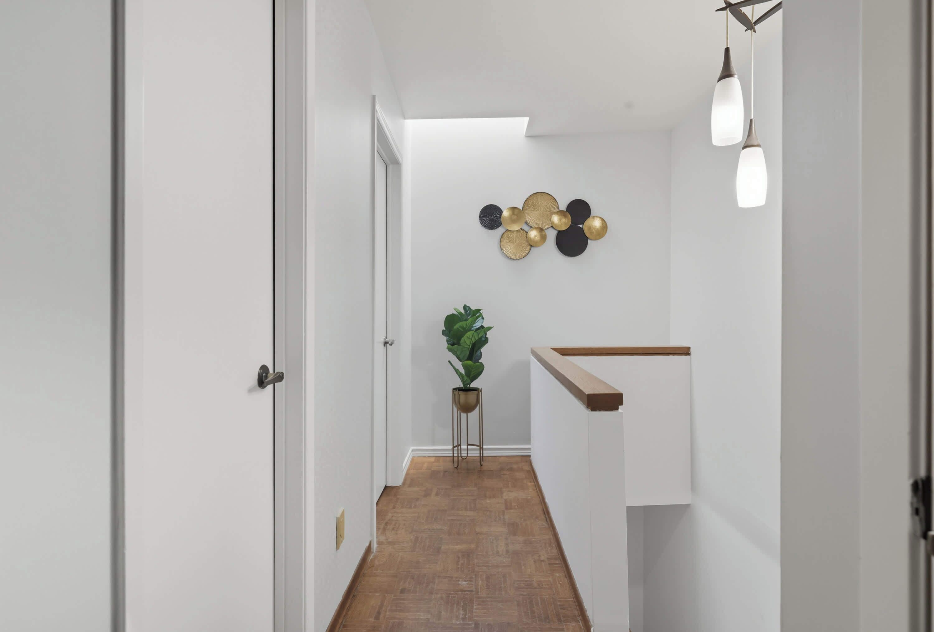 A hallway with a wooden floor, white walls, a plant, and a wall-mounted light fixture.