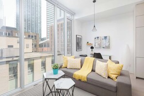 Ein modernes Wohnzimmer mit einem grauen Sofa, gelben Kissen, einem kleinen Tisch mit Pflanze und Blick auf Stadtgebäude durch große Fenster.