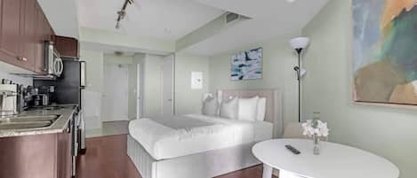 A compact hotel room with a kitchenette, a bed, a bedside table, and a painting on the wall.