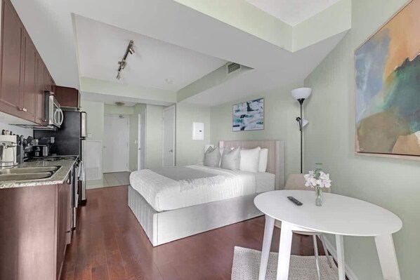 A compact hotel room with a kitchenette, a bed, a bedside table, and a painting on the wall.