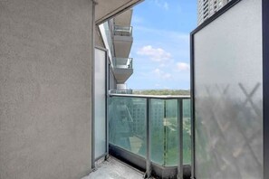 A balcony with a glass railing, a concrete wall, and a view of a building and greenery.
