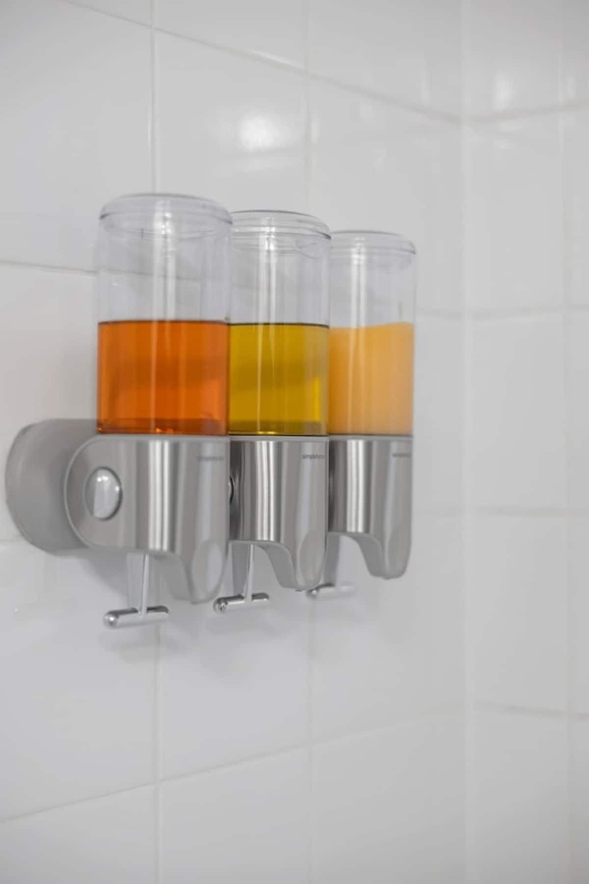 A wall-mounted dispenser with three transparent containers holding orange liquid.